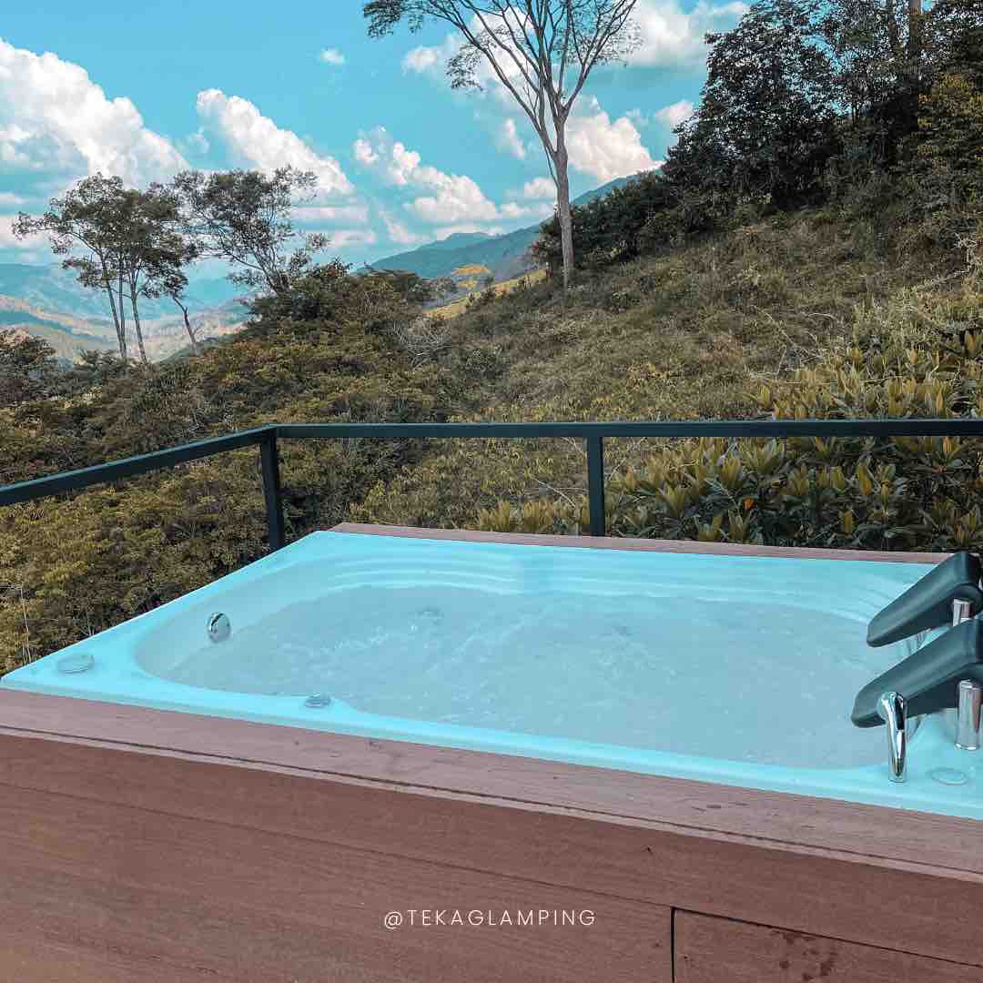 A hot tub is positioned on a wooden deck, overlooking lush green hills and foliage. The clear blue sky is dotted with fluffy clouds, enhancing the serene landscape surrounding the area. The hot tub is filled with water and features stylish modern fixtures.