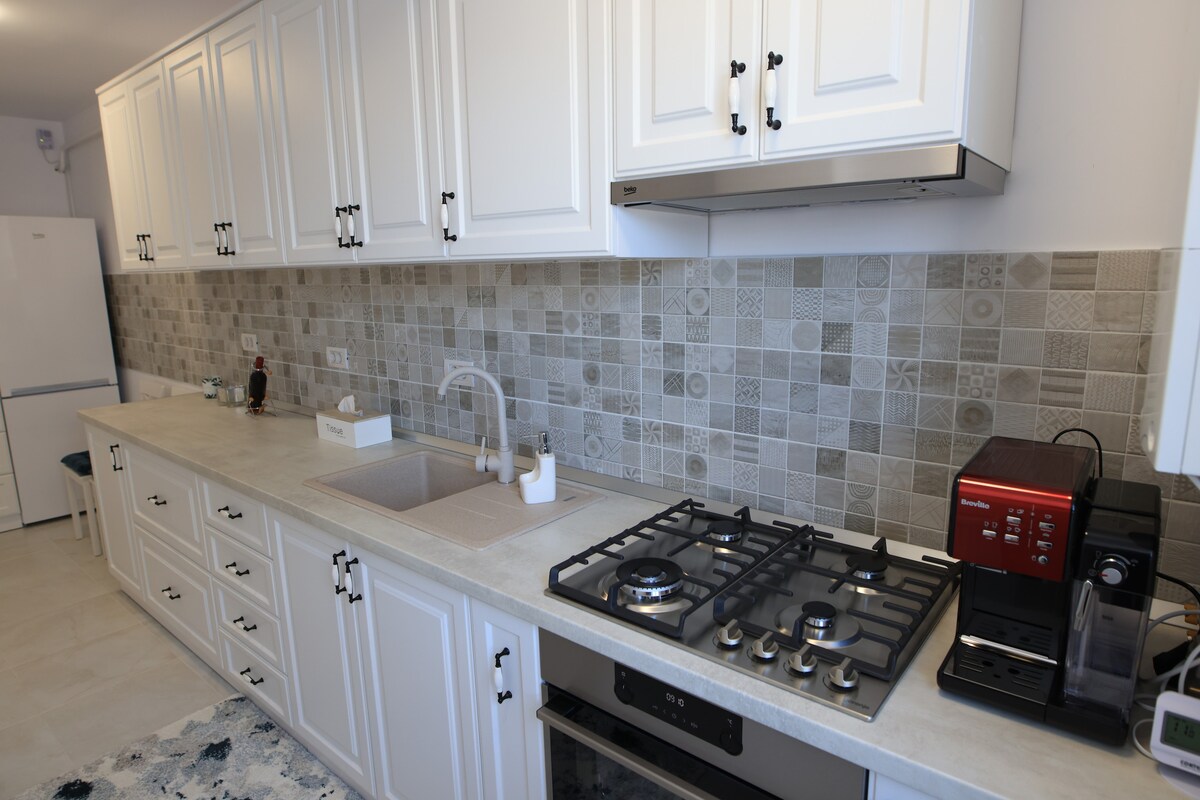 A modern kitchen is presented, featuring white cabinetry, a tiled backsplash with intricate patterns, and a variety of appliances. A gas stove and coffee maker are visible, along with a sink set into the countertop. The layout promotes a functional cooking environment.