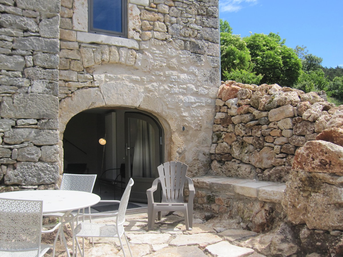 An outdoor area features a round table with four metal chairs and a relaxed Adirondack chair. The weathered stone wall provides rustic charm beside smooth, natural rock seating. A large window reveals glimpses of a well-lit interior, surrounded by lush greenery in a bright blue sky.
