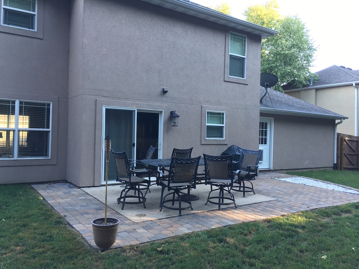 A spacious outdoor patio is equipped with a large table surrounded by six comfortable chairs. A shaded area is created by the overhang of the house. The well-maintained yard features grass and a stone pathway leading to the patio area, offering ample space for gatherings.