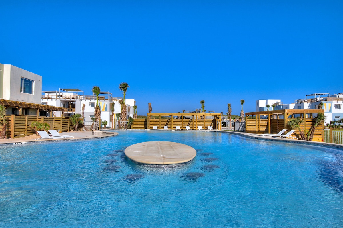 A large outdoor swimming pool is surrounded by sun loungers and palm trees. A circular platform is situated in the center of the pool, reflecting the clear blue sky. The modern resort buildings are visible in the background, providing a relaxing environment.