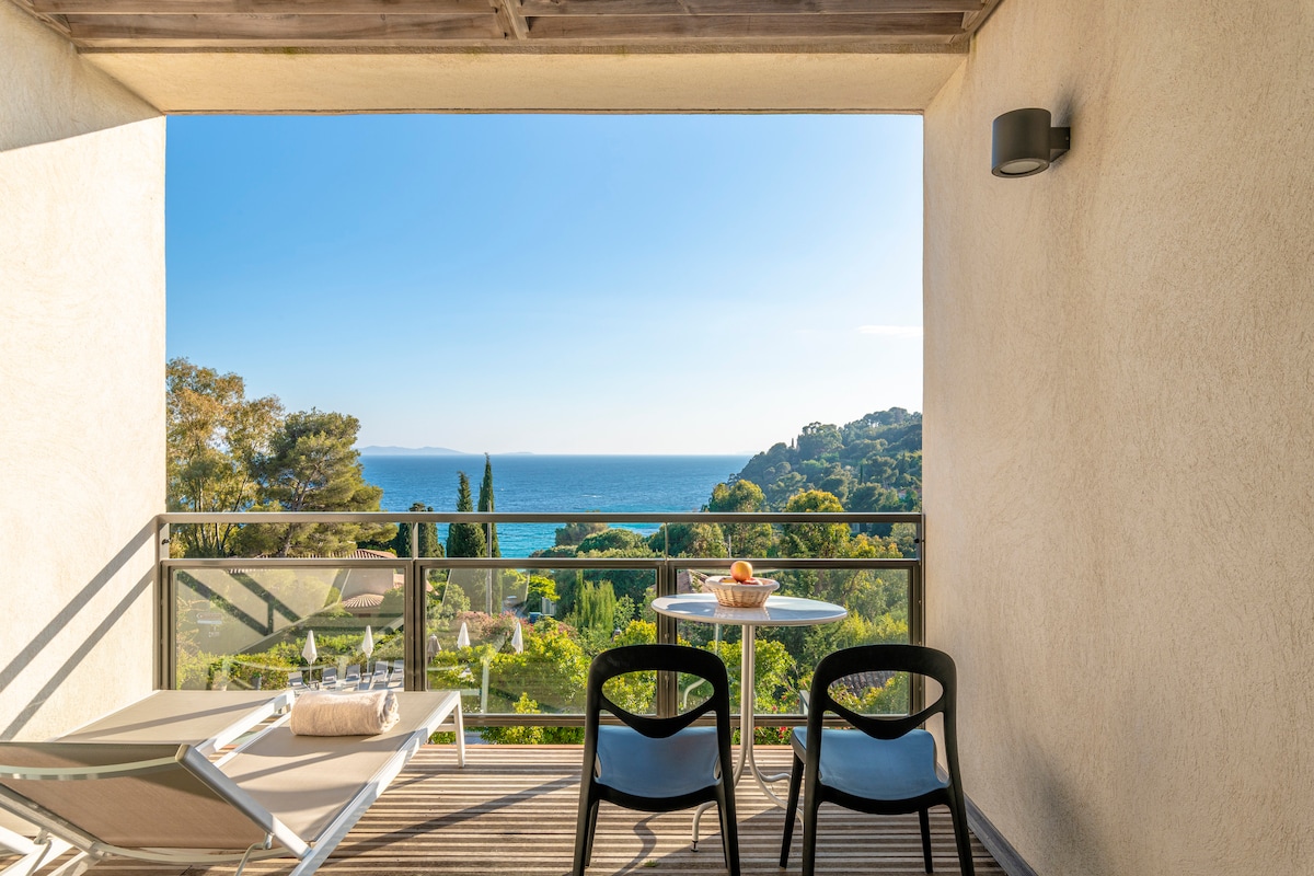 A spacious balcony is equipped with two loungers, a table, and two chairs, offering a clear view of the Mediterranean Sea. Lush greenery is visible in the foreground, while light fills the space from the bright, sunny sky.