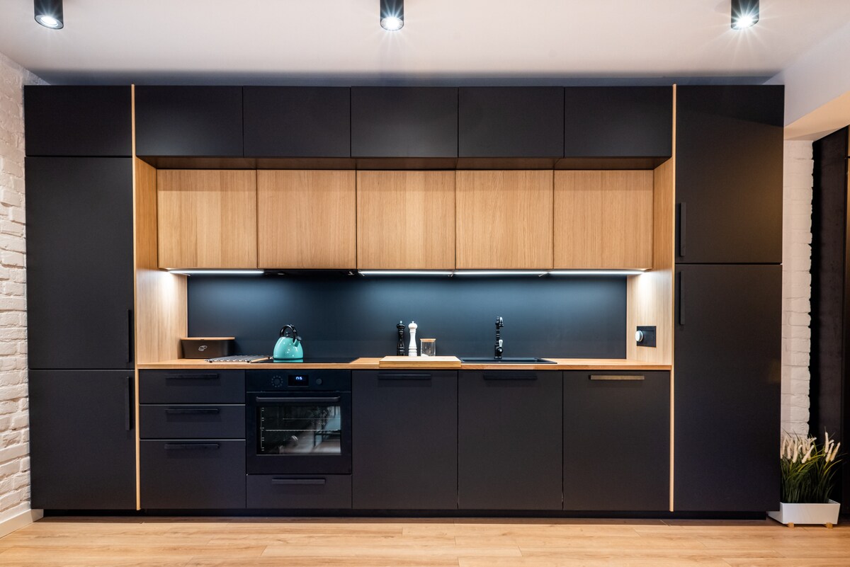A modern kitchen features dark cabinetry paired with wooden accents. Counter space is available under overhead storage, and appliances include an oven and a kettle. Soft lighting highlights the sleek design, creating an inviting working area for meal preparation.
