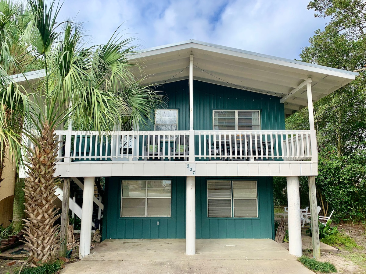 The exterior of the duplex is highlighted with a teal facade and a wide front deck supported by white pillars. Lush palm trees frame the entrance, while large windows allow natural light into the upper unit. The driveway below offers parking space in front of the house.