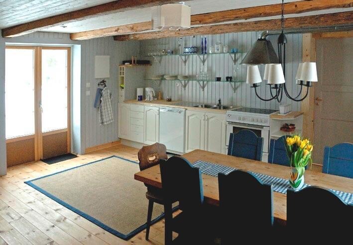 A spacious kitchen and dining area features wooden beams across the ceiling and light grey paneling on the walls. A large wooden dining table is surrounded by black chairs, with a vase of fresh tulips as a centerpiece. The kitchen cabinets are stocked with dishware above a modern stove.