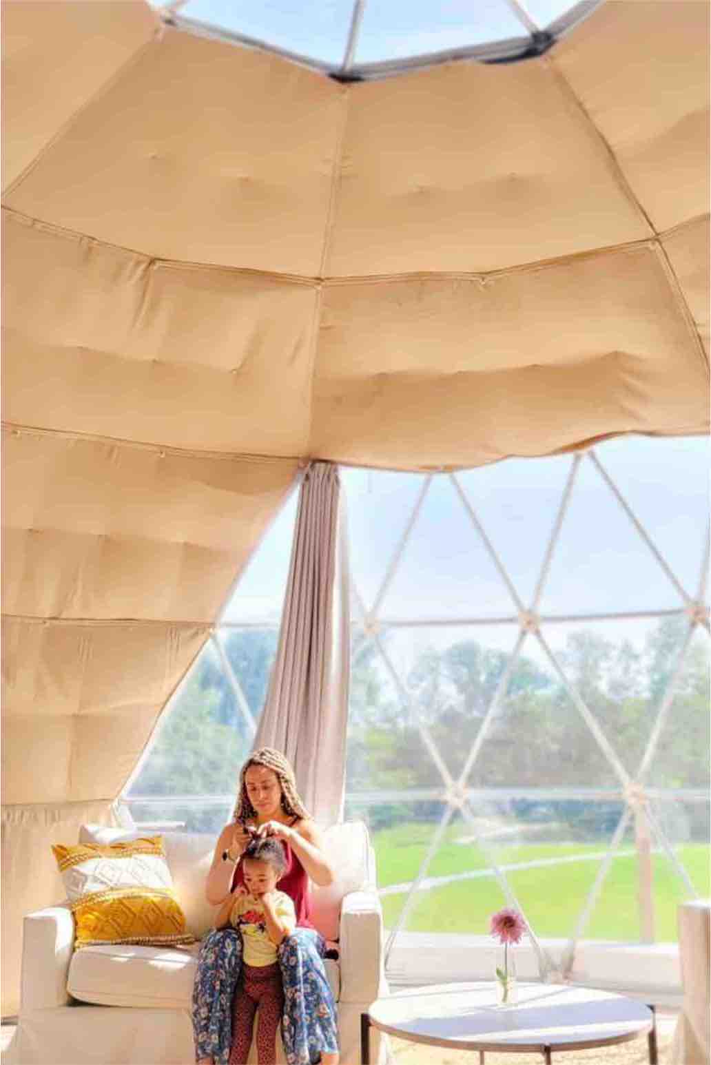 A cozy seating area is showcased in a geodesic dome, featuring a light-colored armchair with a decorative pillow. Natural light streams through large transparent panels, creating an airy atmosphere. A small round table sits nearby, accompanied by a single flower in a vase.