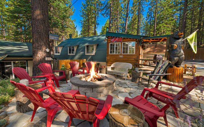 A stone fire pit is surrounded by multiple red Adirondack chairs, set in a natural outdoor area filled with trees. A wooden cabin can be seen in the background, complemented by a carved bear statue, creating a rustic gathering space.