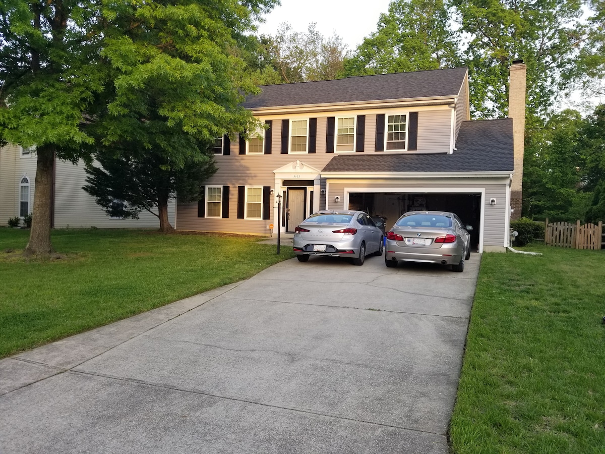 A two-story house is set on a grassy lawn, featuring a driveway with two parked vehicles. The exterior showcases a combination of light siding and dark shutters, with a welcoming front porch and tall trees offering shade.