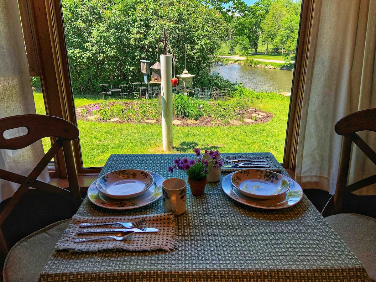 A dining table is set with plates, utensils, and a small potted plant. The large window overlooks a lush green garden and the Iowa River, with a bird feeder visible in the background. Natural light brightens the inviting space.