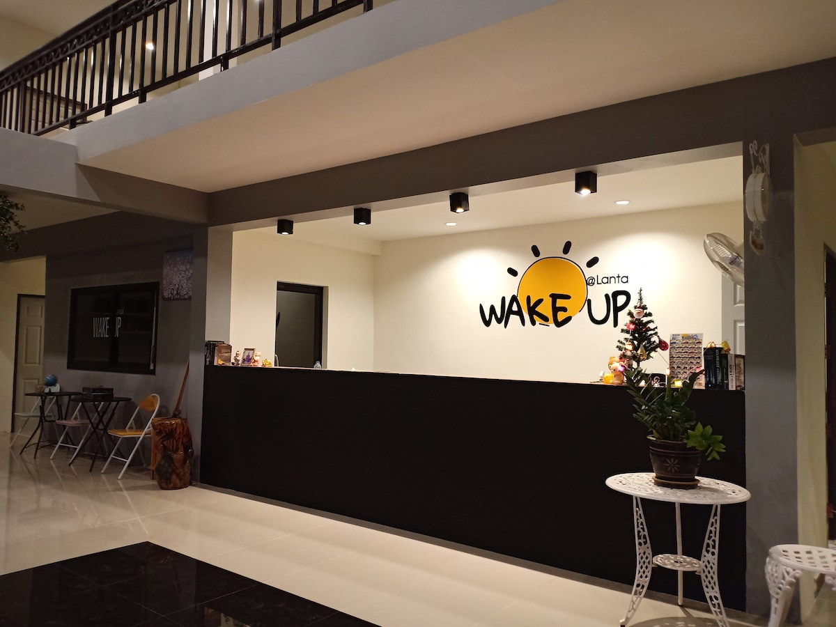 A reception area is provided with a modern counter featuring the 'WAKE UP' logo. Adjacent seating options include folding chairs and a small round table. A decorative plant adds a touch of greenery, while warm lighting creates an inviting space for guests.