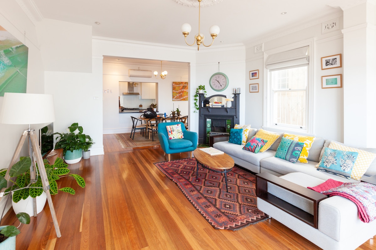 The living area features polished wooden floors and a comfortable sectional sofa adorned with colorful cushions. A vibrant rug sits beneath a central wooden coffee table. Light from large windows highlights a green plant and a decorative lamp, while a kitchen area is visible in the background.