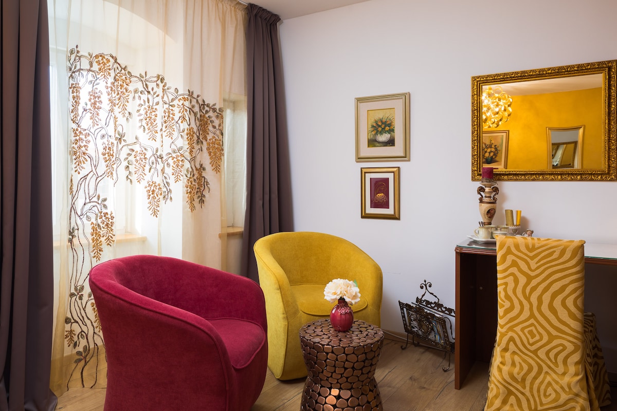 A well-arranged sitting area features two accent chairs in yellow and red, positioned beside a small, circular table with a metallic finish. A decorative plant sits on the table, while curtains with floral patterns frame the window. A mirror and artwork adorn the walls.