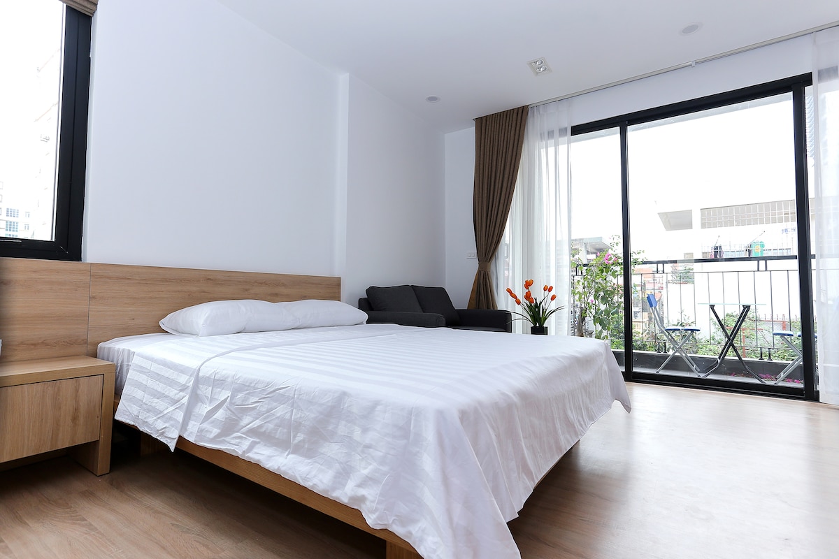 A spacious room features a large bed with a crisp white bedspread. Natural light floods the area through a floor-to-ceiling window, which opens onto a small balcony with outdoor seating. A comfortable chair adds a relaxing touch to the modern decor.