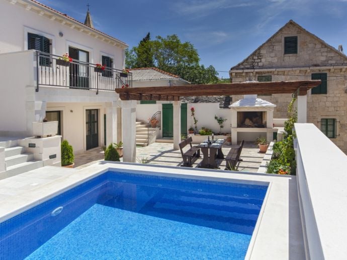 The outdoor area features a private pool surrounded by a stone terrace. A wooden pergola provides shade over dining furniture. Potted plants add greenery, and a stone barbecue is visible nearby. The surrounding stone buildings reflect traditional architecture, creating a harmonious outdoor setting.