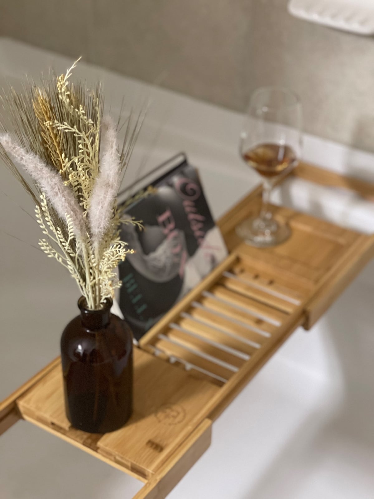 An elegant bamboo bath tray rests across the bathtub, featuring a small dark glass vase filled with dried flowers. A glass of wine and a magazine are positioned on the tray, creating a serene atmosphere for relaxation.