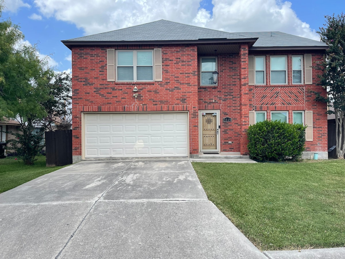 The exterior of a two-story brick house is presented, featuring a spacious driveway leading to a double garage. Lush green grass frames the front yard, complemented by neatly trimmed shrubs. Large windows adorn the upper and lower levels, allowing for natural light to enter.