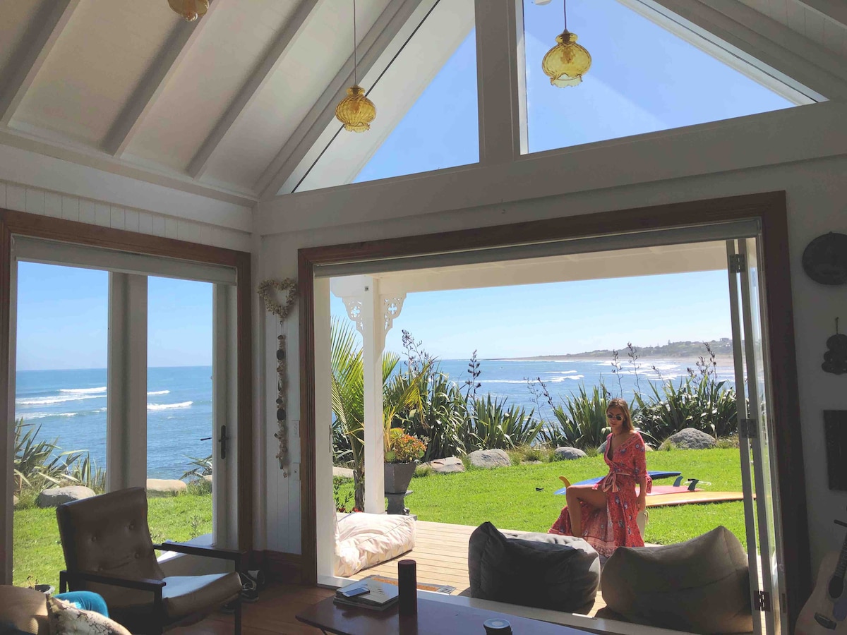Large windows allow ample natural light to fill the interior, offering unobstructed views of the ocean. A spacious deck is visible outside, framed by lush greenery. Comfortable seating arrangements, including armchairs, invite relaxation.
