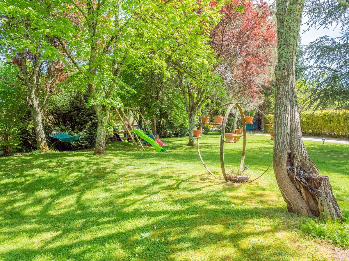 A lush garden area is captured, featuring vibrant green grass and a mix of deciduous trees. A garden slide and swing set are positioned under the trees, while a hammock is seen draped between two branches, providing a relaxing outdoor space.