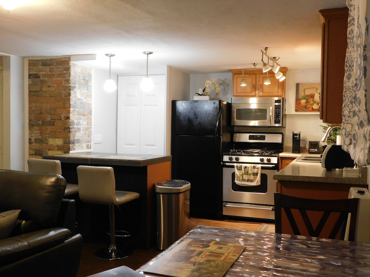 An open kitchen area features modern stainless steel appliances, including a refrigerator and stove. A breakfast bar with two upholstered stools is positioned by a stone accent wall, while overhead lights provide a warm glow. A dining table is visible in the foreground.