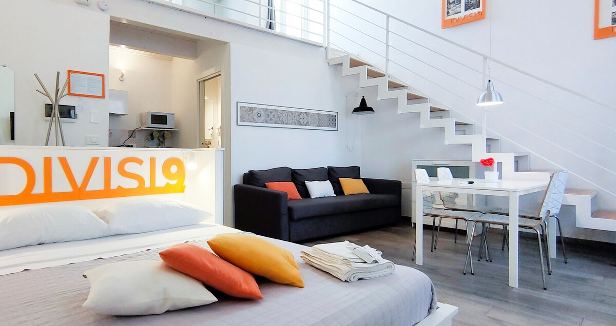 The vibrant living area includes a spacious bed with colorful pillows, a comfortable sofa, and a dining table set for four. The open layout is accentuated by modern furnishings and a staircase leading to an upper level. Natural light fills the space through large windows.