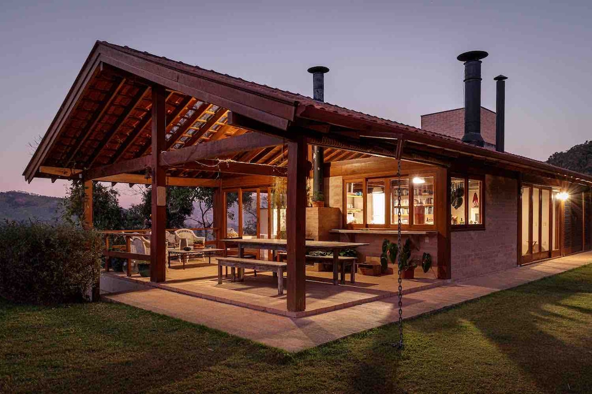 A contemporary vacation home features a spacious covered patio with wooden beams, showcasing seating areas. Large windows provide views of the surrounding landscape. The structure is complemented by sloped roofs and chimney stacks, offering a harmonious blend with nature.