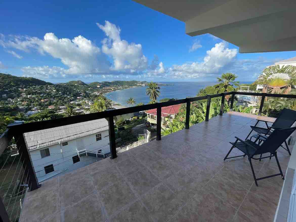 An oversized balcony offers stunning views of the coastline and surrounding landscape. Sunlight fills the space, highlighting the spacious tiled flooring. Two lounge chairs are positioned for relaxation, with a railing providing safety while allowing unobstructed sightlines of the beach and hills beyond.