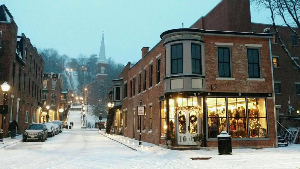 A charming street scene captures a snow-covered downtown area at dusk. Brick buildings, including the Simply Elegant Boutique, are illuminated with warm light. A gentle slope is visible in the background, leading to a steeple that tops the hill, surrounded by falling snowflakes.
