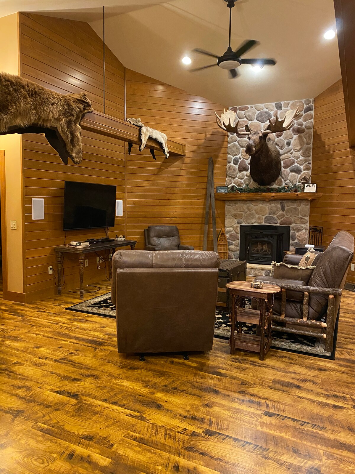 A rustic living area features wooden walls and a stone fireplace. Taxidermy mounts of a moose and smaller animals are displayed overhead. Two leather chairs are positioned near the fireplace, with a wooden side table beside one. A television is mounted on the wall.