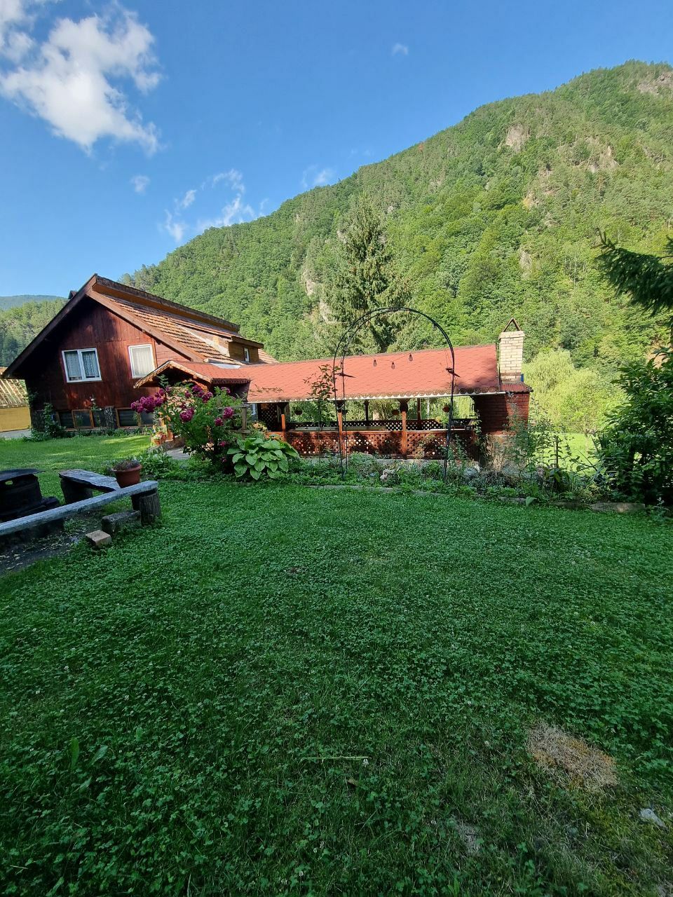 A charming wooden cabin sits nestled against a lush green hillside, with a red roof and surrounding gardens. The well-maintained lawn features colorful flowers and plants, contributing to a serene outdoor environment. The mountainside serves as a picturesque backdrop.