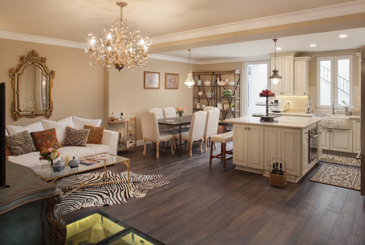 A spacious living area features a comfortable white sofa adorned with decorative pillows, paired with a glass-top coffee table. A dining table with upholstered chairs is positioned nearby, complemented by warm lighting from stylish fixtures. The kitchen, with its modern cabinetry, showcases an island with seating.