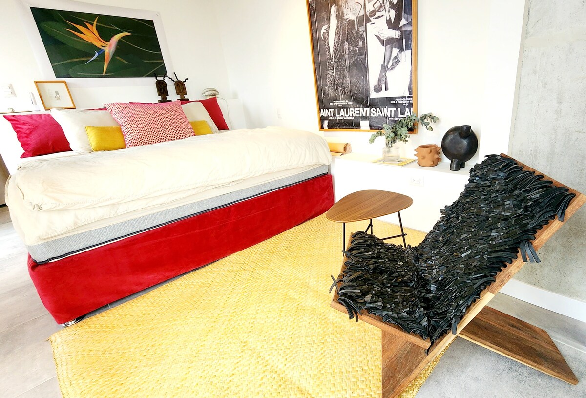 A spacious sleeping area features a elevated bed with a red base, layered with warm linens and decorative pillows. A unique lounge chair, crafted from natural materials, complements the space. A small wooden table sits nearby, with textured flooring enhancing the overall aesthetic.