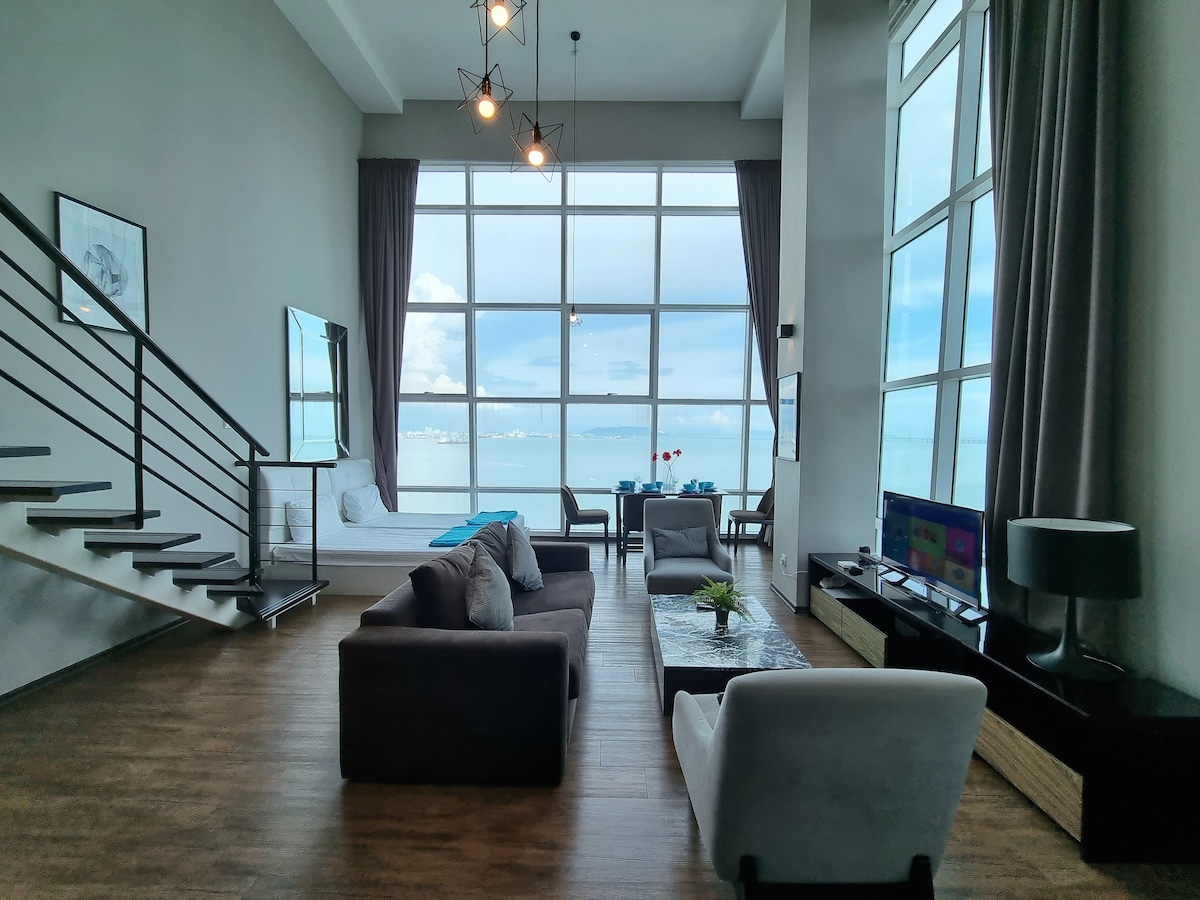 A spacious living area is showcased, featuring large windows that provide a panoramic view of the sea. The area includes a comfortable seating arrangement with a sofa and chairs, alongside a coffee table. A staircase leads to an upper level, while a cozy bed is visible in the background.