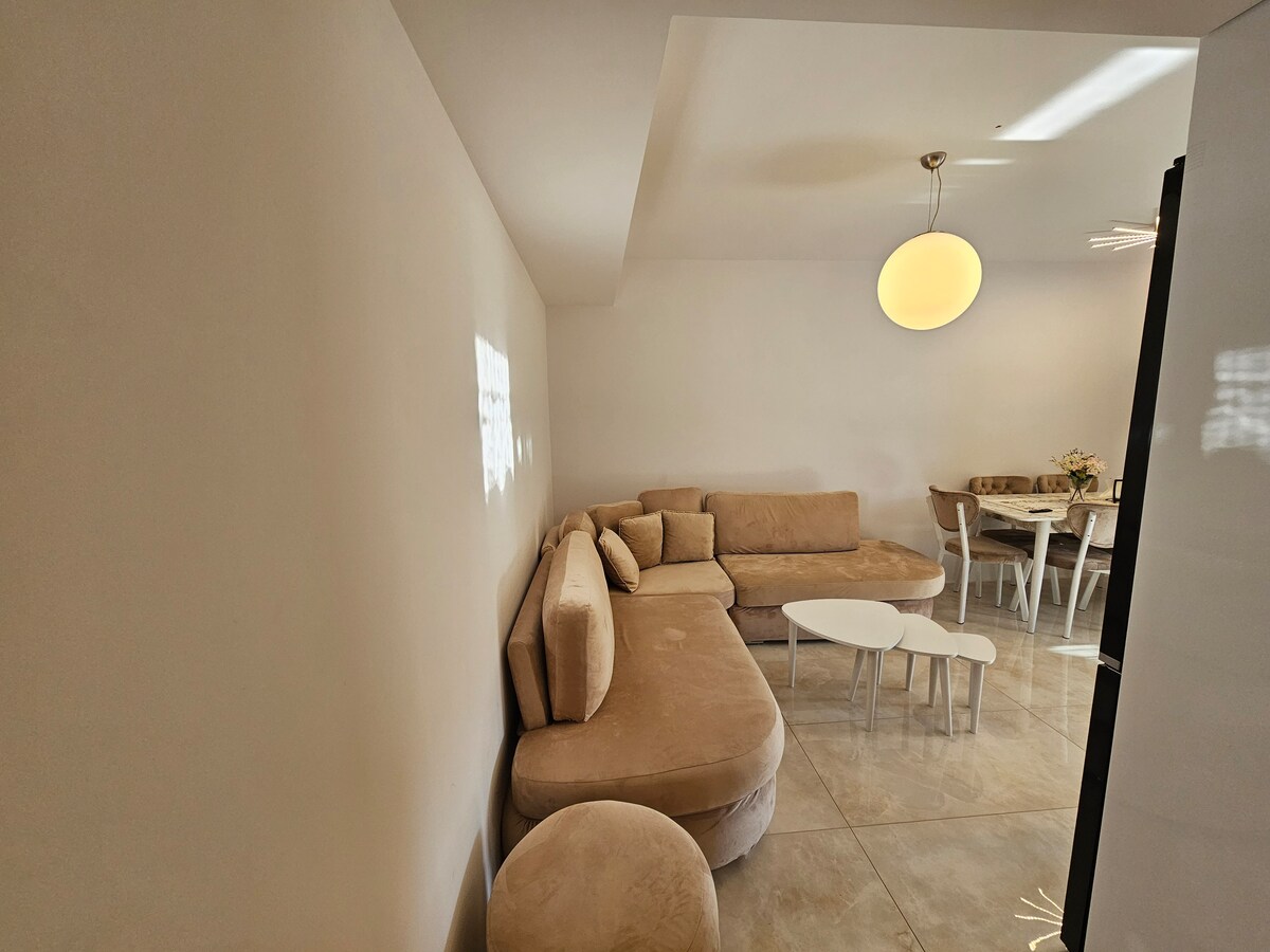 A light-filled living area features a soft, neutral-toned sectional sofa arranged around a small coffee table. A round ceiling light provides illumination. In the background, a dining table with chairs is visible, enhancing the inviting layout of the space.