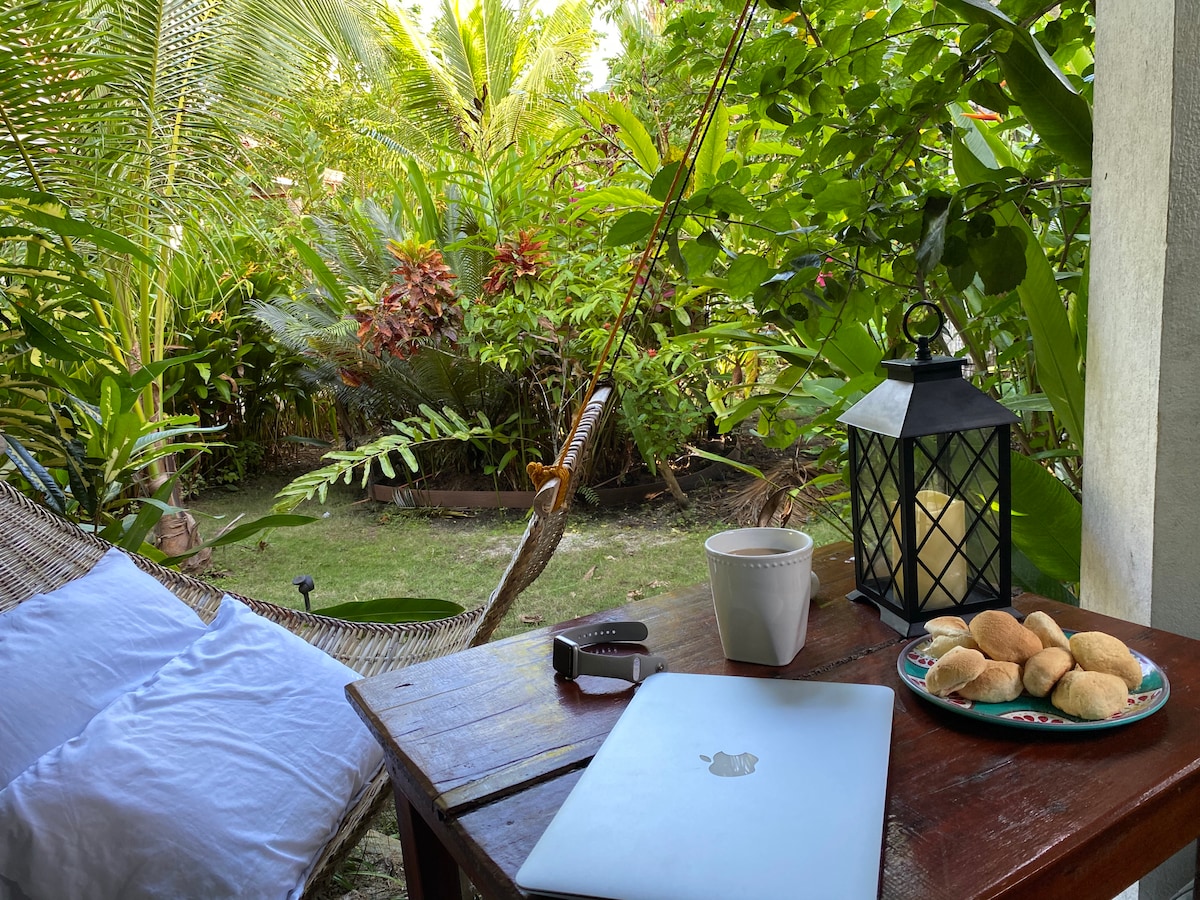 A wooden table is set with a laptop and a plate of cookies beside a white mug, suggesting a relaxed workspace. A lantern adds subtle light, while a hammock drapes nearby, framed by lush greenery and colorful plants in the background.