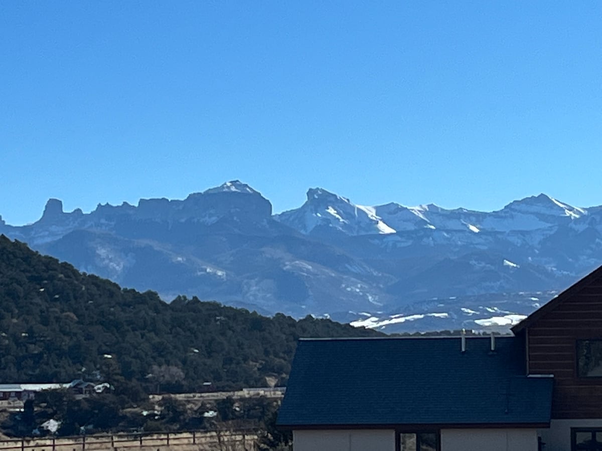 Snow-capped mountains rise in the distance against a clear blue sky. A gently sloping landscape of trees and fields is visible in the foreground, leading up to the mountainous horizon. The scene captures the natural beauty surrounding the area, providing a serene backdrop.