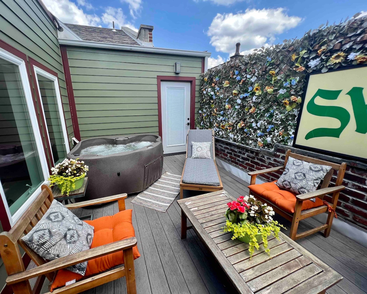 An enclosed private patio features a two-person hot tub and wooden seating. Brightly colored flowers adorn the tables, and a rug enhances the space. Walls are decorated with floral accents, creating a serene outdoor environment perfect for relaxation.