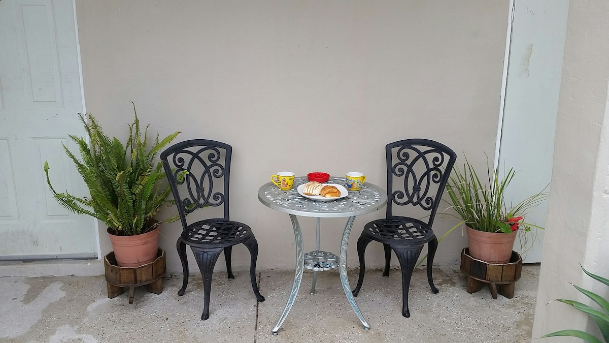 A small outdoor seating area features a round table with two decorative chairs. Two cups and a small plate of pastries are placed on the table. Potted plants flank the seating area, adding natural elements to the space.