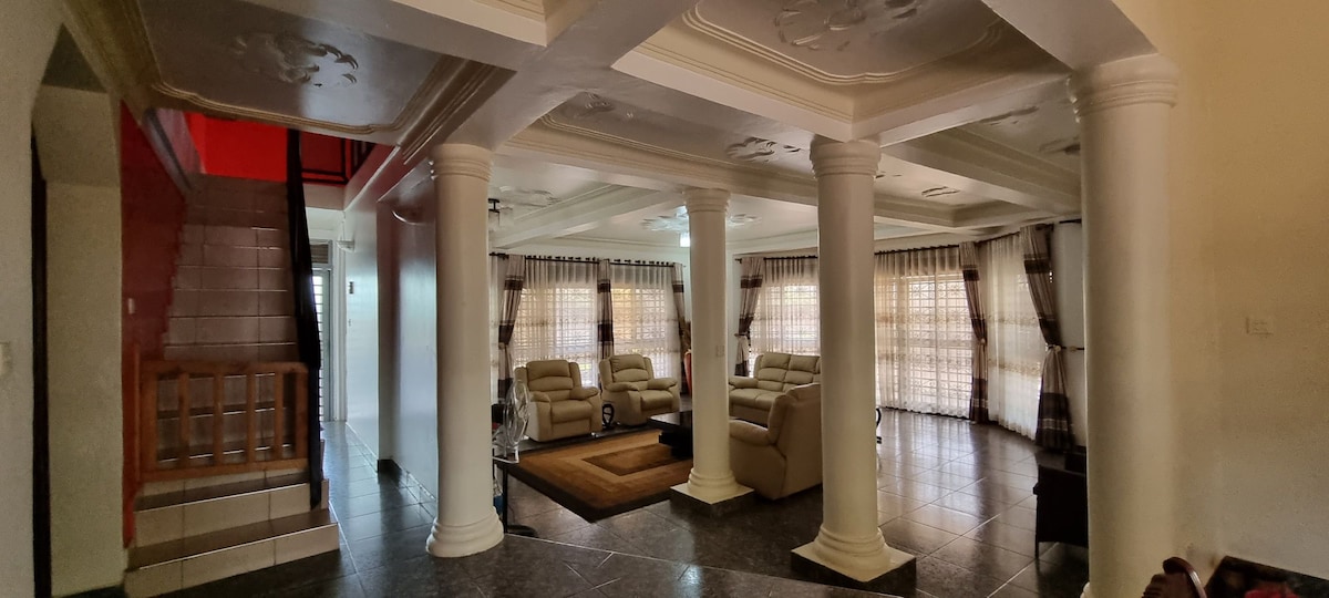A spacious living area features decorative ceiling designs and three large windows dressed with sheer curtains, allowing natural light to fill the room. Two beige sofas are arranged around a central rug, while tall white columns add elegance to the open layout.