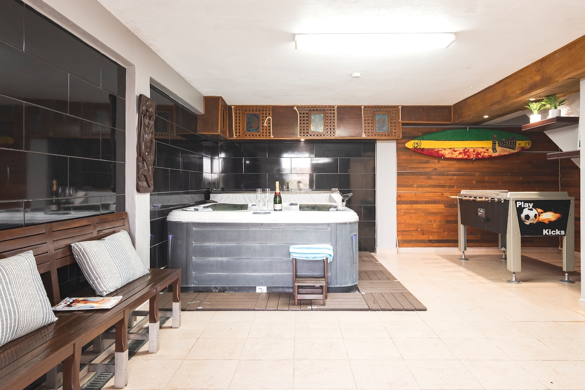 A private jacuzzi comfortably accommodates several guests, surrounded by modern wooden paneling and dark tiles. Nearby, a foosball table and seating area featuring wooden benches with cushions provide a relaxed space for entertainment and socializing.