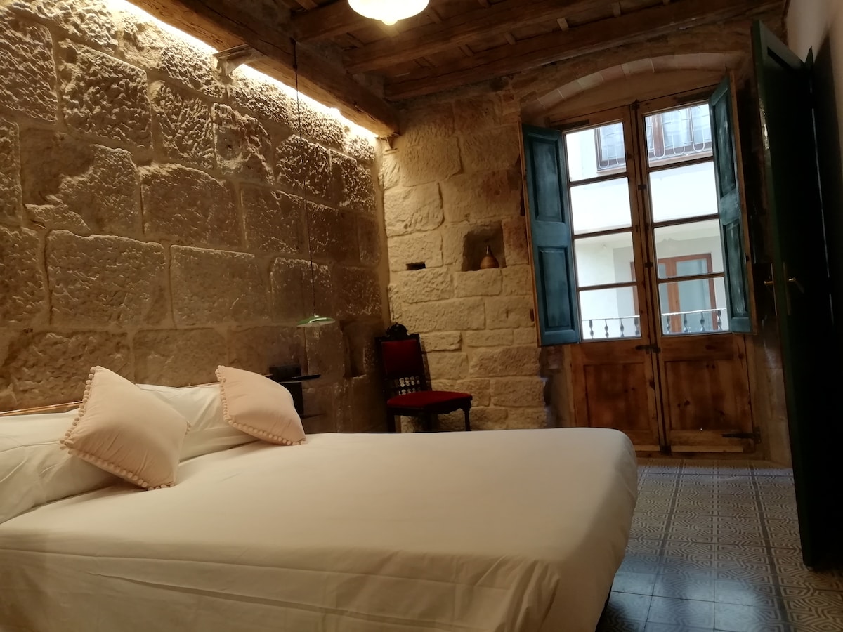 A double bedroom is presented with a spacious bed featuring soft pillows. Exposed stone walls retain historical charm, complemented by decorative tiles on the floor. Large windows with wooden shutters allow natural light, while a single chair adds to the room's decor.