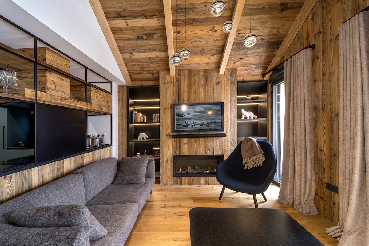 A modern living area is presented with wooden walls and ceiling beams. A grey sofa is arranged next to a black coffee table. A large television is mounted on the wall, surrounded by black shelving with decorative items. Natural light floods in through the windows, which are dressed with soft curtains.