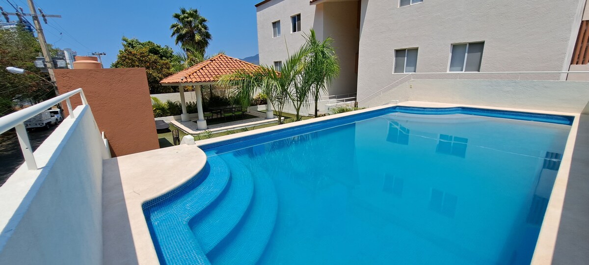 A clear blue swimming pool is framed by a well-maintained patio area. Green palm trees provide shade, while a gazebo is visible in the background. The building's light exterior complements the inviting atmosphere of this outdoor space.