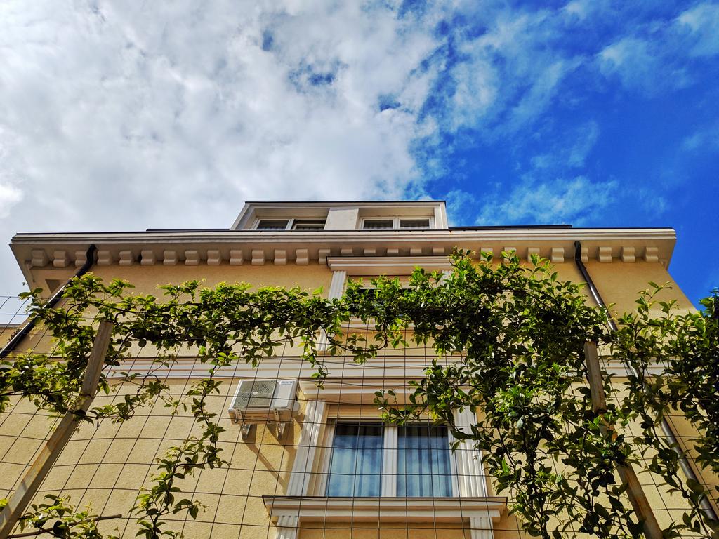 A view of the building’s exterior is captured from below, displaying a light-colored facade adorned with greenery. A clear blue sky enhances the scene, complemented by fluffy clouds. The architectural details are visible, including air conditioning units and large windows.