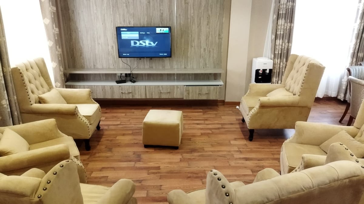 A cozy lounge area is arranged with four beige armchairs facing a mounted TV. A small ottoman is situated in the center, and light hardwood flooring complements the neutral tones. Soft curtains frame the large windows, allowing natural light to gently illuminate the space.