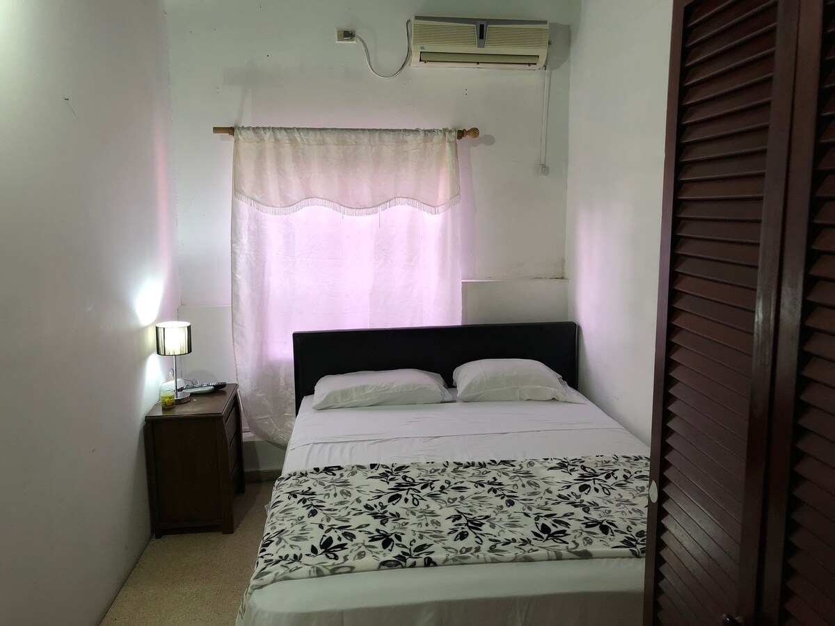 A cozy bedroom is fitted with a spacious bed adorned with a floral bedspread. Natural light filters through a sheer curtain, softly illuminating the room. A bedside table with a lamp is positioned next to the bed, and an air conditioning unit is mounted on the wall.