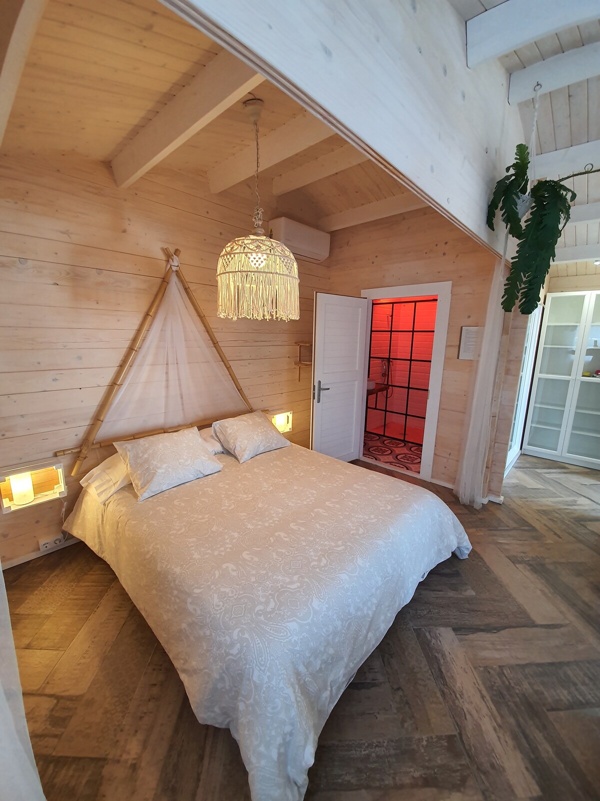 A cozy sleeping area features a spacious bed with soft linens and decorative pillows. Warm wooden walls create a natural ambiance, complemented by a charming hanging light fixture. A doorway leads to the bathroom, while light enters through sheer curtains.