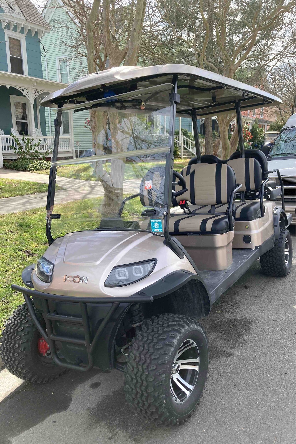 A six-passenger golf cart is shown parked along a residential street. It features a sturdy frame with black wheels and a canopy over the seating area. The seats are cushioned in cream and dark accents. Nearby, trees and historical homes are visible, providing a quaint neighborhood backdrop.
