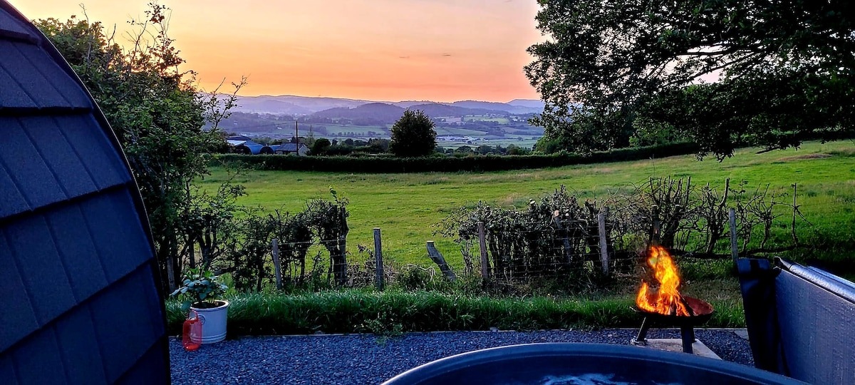 The image captures a scenic view at sunset, featuring rolling hills and distant mountains. A firepit with flames is visible in the foreground, surrounded by greenery. The gentle glow of the sunset casts a warm light across the landscape, enhancing the serene atmosphere.