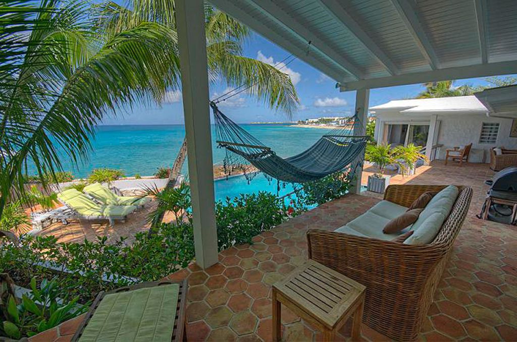 A spacious patio is surrounded by lush tropical vegetation, featuring a hammock strung between palm trees. A swimming pool is visible in the foreground, blending into the sea beyond. Comfortable seating arrangements are present, offering a relaxed atmosphere with clear ocean views.