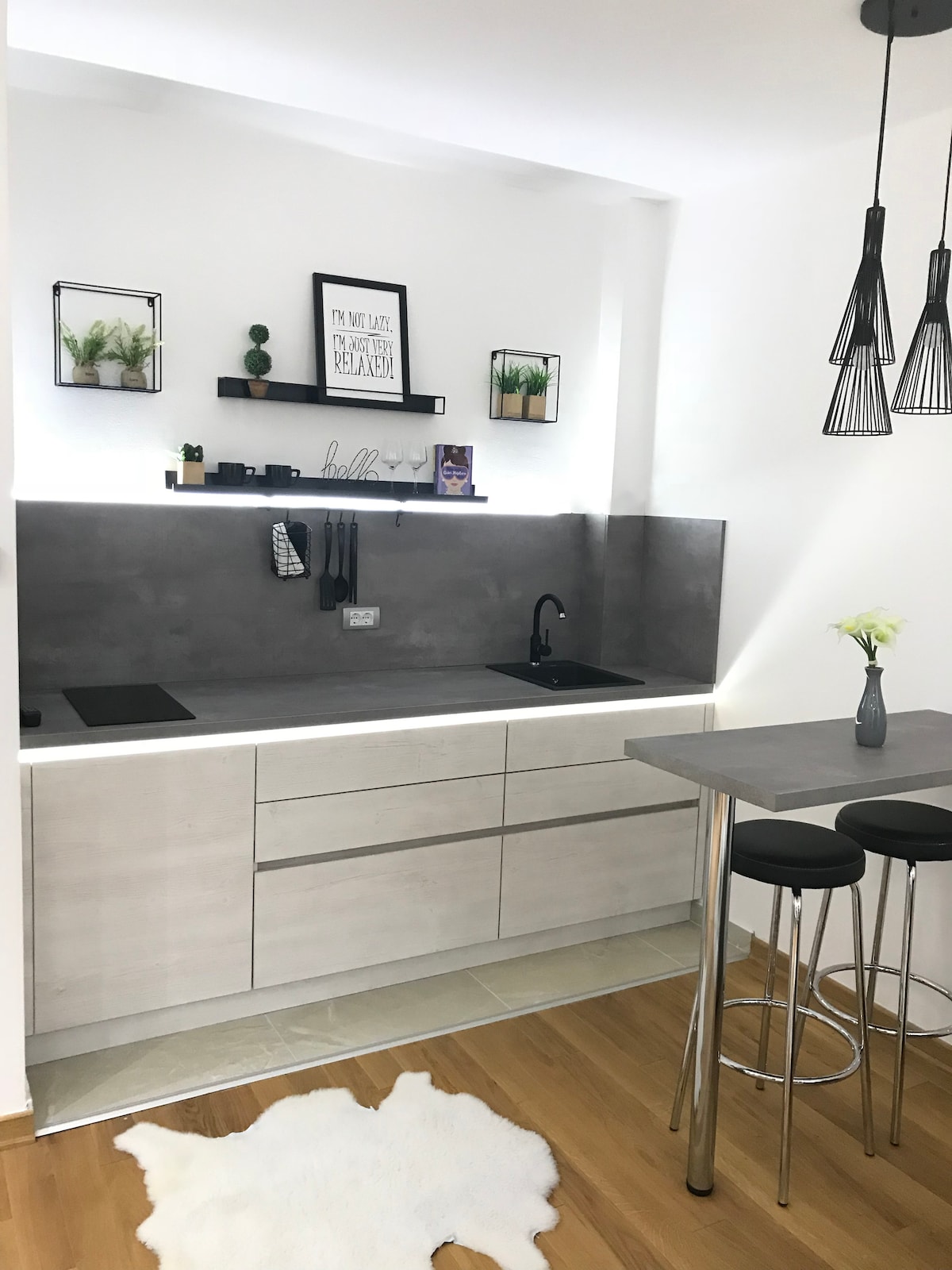 A modern kitchen is displayed, featuring clean lines and a minimalist design. Sleek cupboards provide ample storage, while a stylish counter offers seating for two. Subtle lighting highlights the integrated sink and cooking area, complemented by decorative plants and wall art above.
